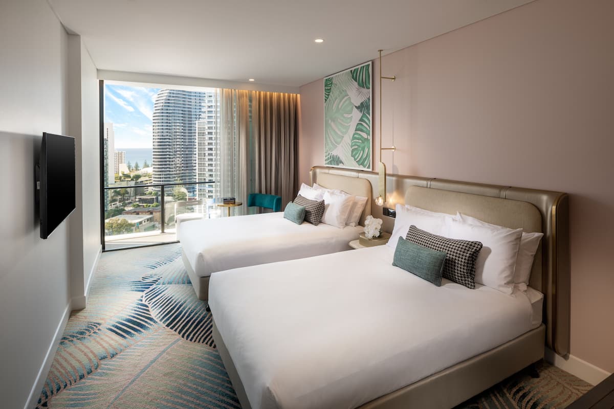 A hotel room with two beds, a large window with a city view, a flat-screen TV, and a patterned carpet.
