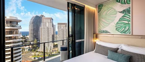 A hotel room with a balcony, a bed with multiple pillows, a large window with a city view, and a tropical leaf painting on the wall.