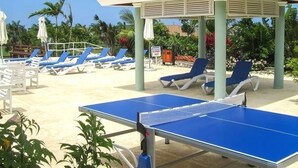 Un área de piscina con sillones reclinables azules, una mesa de ping pong y un pabellón.