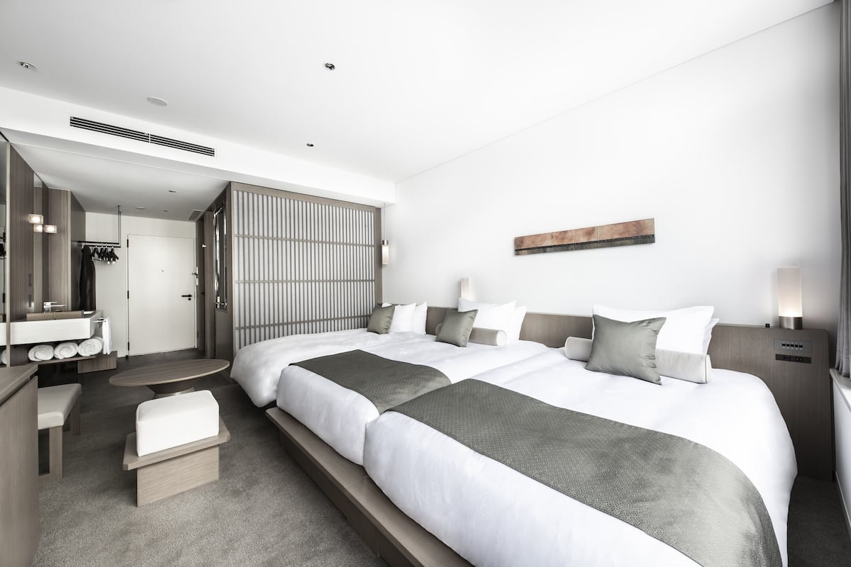 A modern hotel room with two beds, a sitting area, a vanity, and a large window.