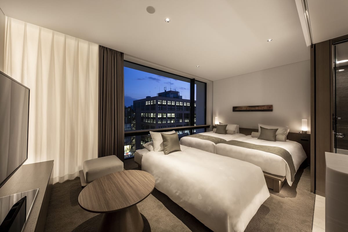 A modern hotel room with two beds, a TV, and a large window with a city view.