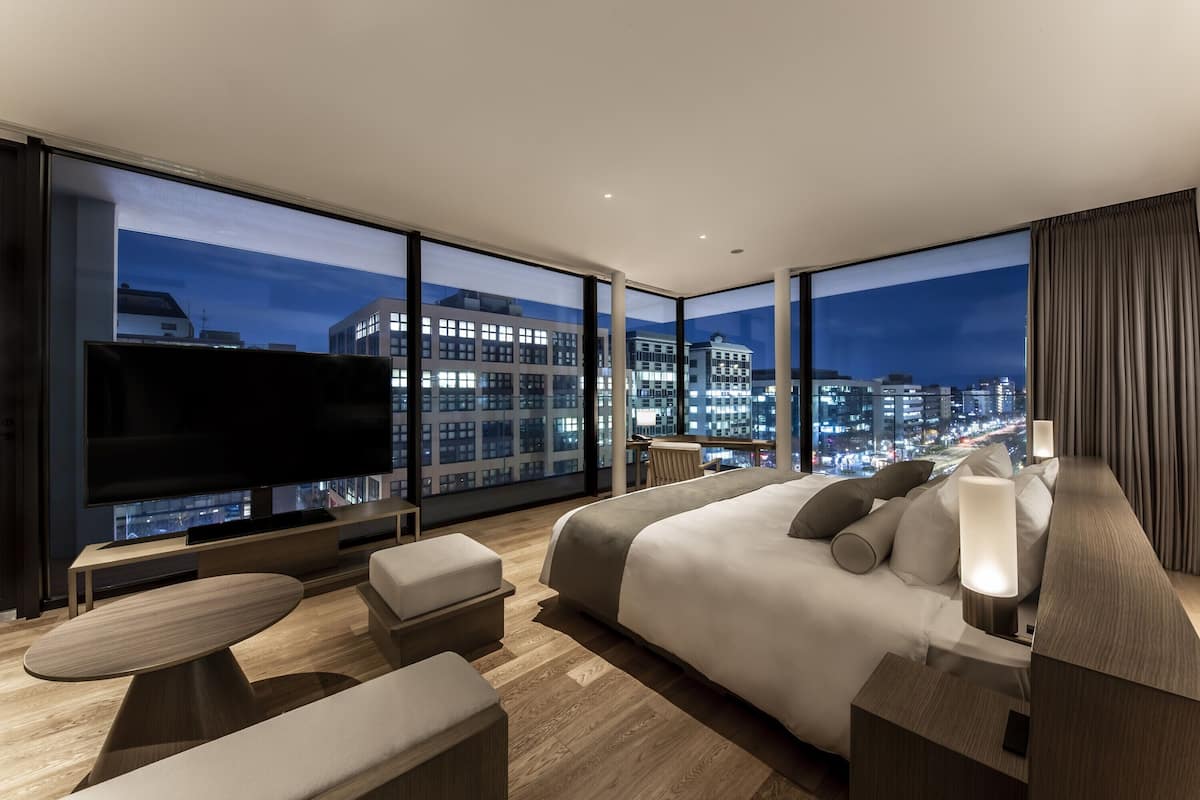 A modern hotel room with a large bed, a flat-screen TV, a dining area, and a city view.