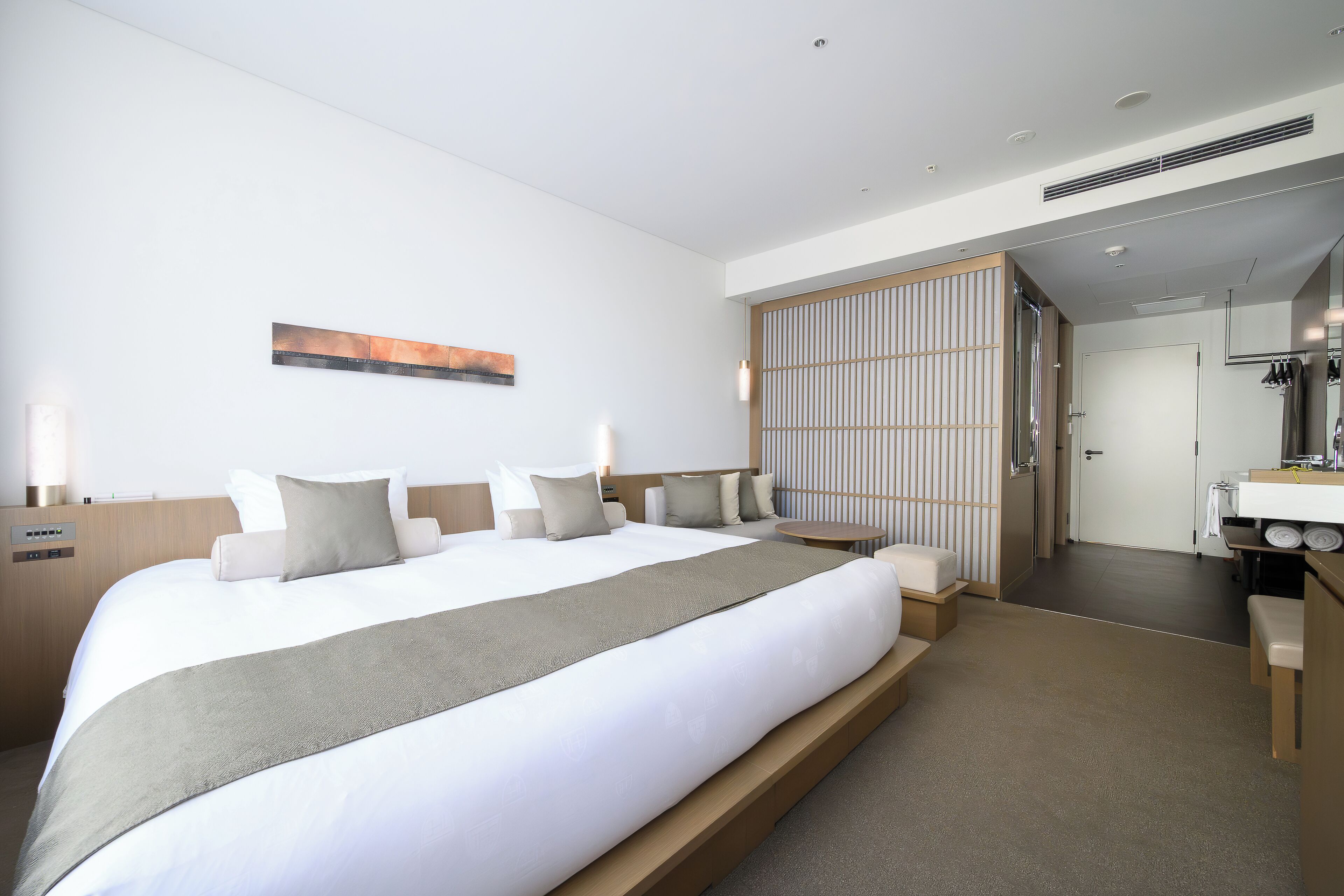 Una habitación de hotel moderna con una cama grande, mesitas de noche, una pequeña mesa redonda, un baño con espejo y un armario con puerta corredera.