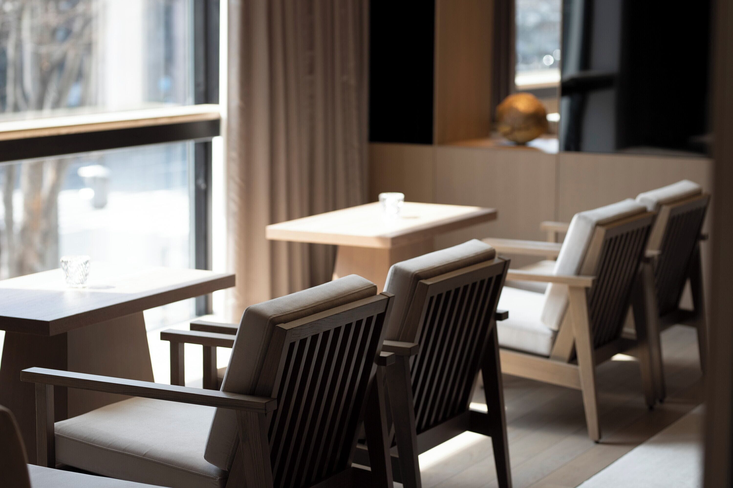 Et moderne spiseområde med træborde og stole, store vinduer med gardiner, og et minimalistisk indretning.