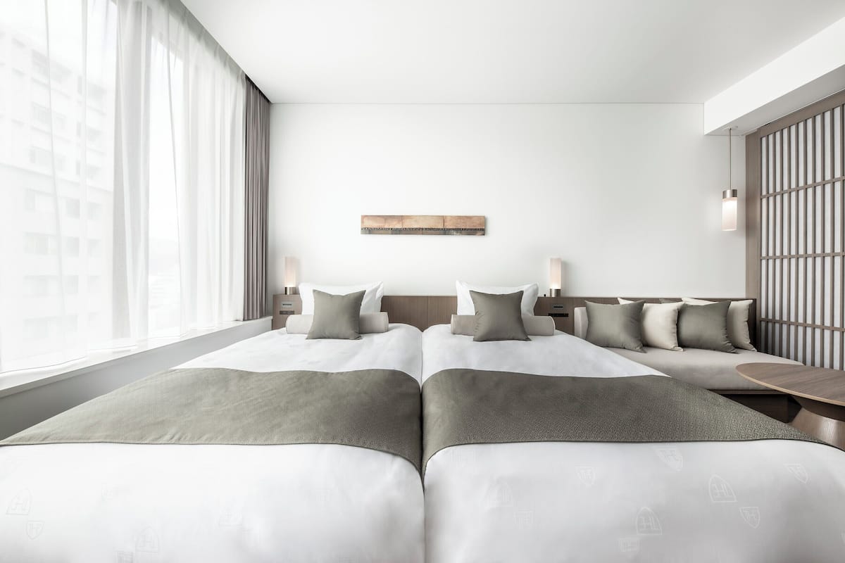 A modern hotel room with two beds, a large window, a wooden headboard, and a wall-mounted artwork.