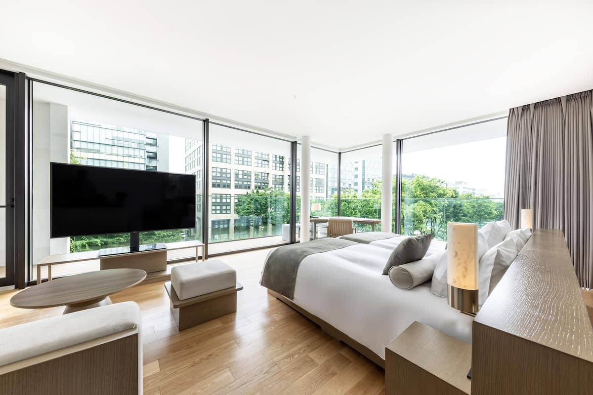 A modern hotel room with a large bed, a flat-screen TV, a seating area, and large windows offering a city view.