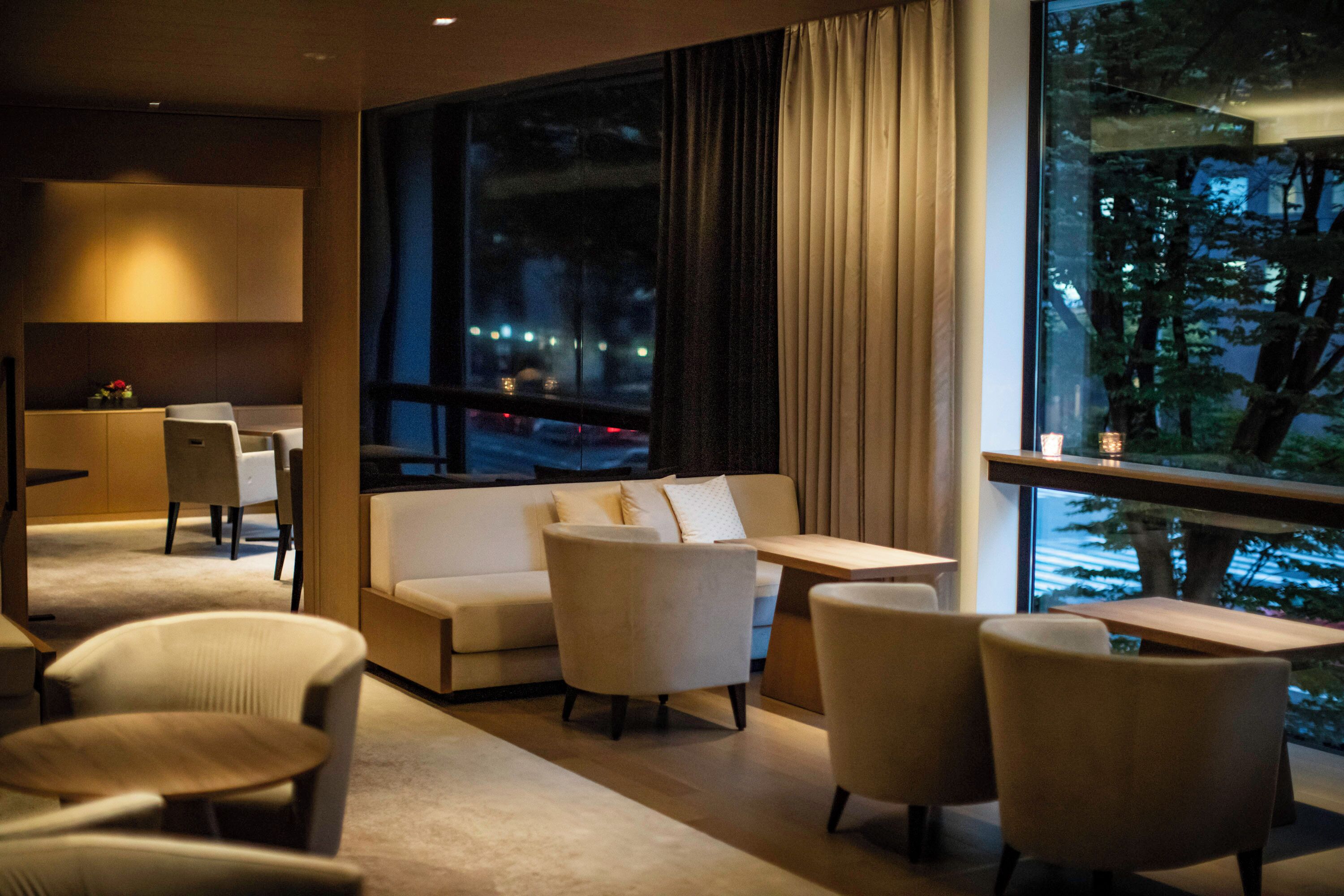 A hotel room with a sofa, chairs, a small table, and a large window with a view of the city at night.