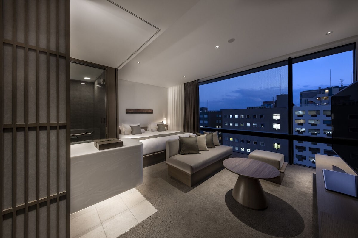 A modern hotel room with a large window, a sofa, a round table, and a small desk area.