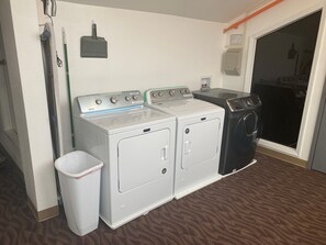 A laundry area with two washing machines and a dryer, a trash bin, and a wall-mounted heater.