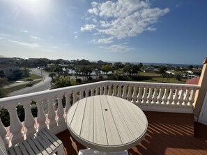 A balcony with a round wooden table and white balustrade, overlooking a coastal view with houses and palm trees. - Waterfront Home - Pool, Boat Dock, Roof Top Patio, Piano, Game Room, Pin Ball (New Port Richey)