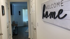 A hallway with a 'Welcome home' sign, a ceiling vent, and a door leading to a living area.