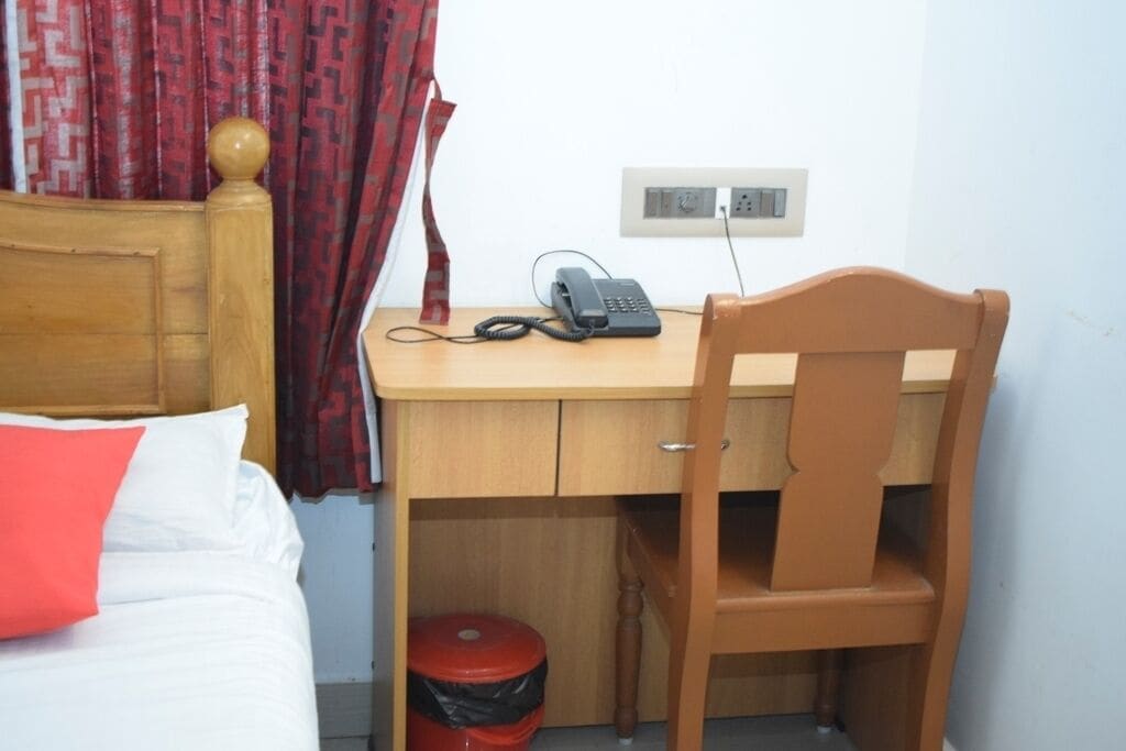 A bedroom with a wooden bed, a desk with a telephone, a red trash bin, and a wall socket.