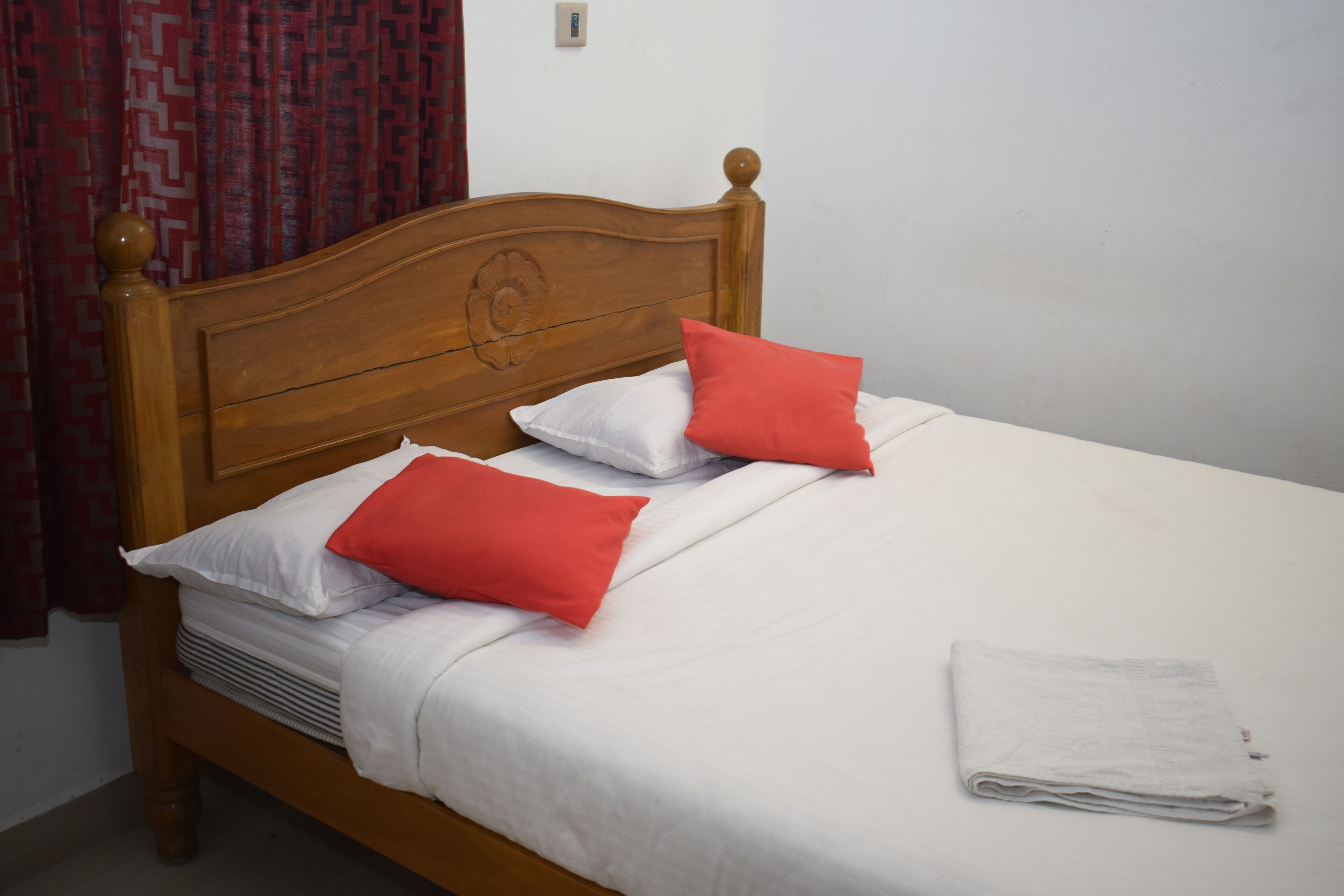 A neatly made bed with white bedding and two red pillows, a wooden headboard, and a folded white towel on the side.