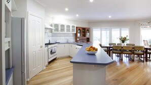 A modern kitchen with white cabinetry, a central island with a bowl of fruit, and a dining area with wooden chairs and a table. - Twin Gums: Large family home, beautiful gardens on banks of Denmark River (Denmark)
