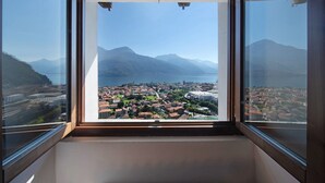 A room with a large window offering a view of a coastal town, mountains, and a body of water. - Vacation Apartment in Beautiful Vineyard 'Crotto Palma' with Lake View & Wi-Fi (Dongo)