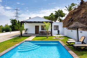 Una piscina con zona de patio, una pequeña casa con área de estar cubierta al aire libre, y una ladera con vegetación.