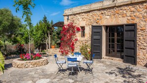 A stone building with a door, a table with chairs, and a garden with flowers. - Pet-Friendly Vacation Home Moli Den Barret with Mountain View, Pool & Wi-Fi (Santa Margalida)