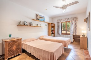 Une chambre avec deux lits, une armoire en bois, une bibliothèque, une fenêtre avec des rideaux et un ventilateur de plafond.