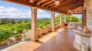 Un patio techado al aire libre con vistas al paisaje, columnas de piedra, plantas en macetas y mobiliario exterior.