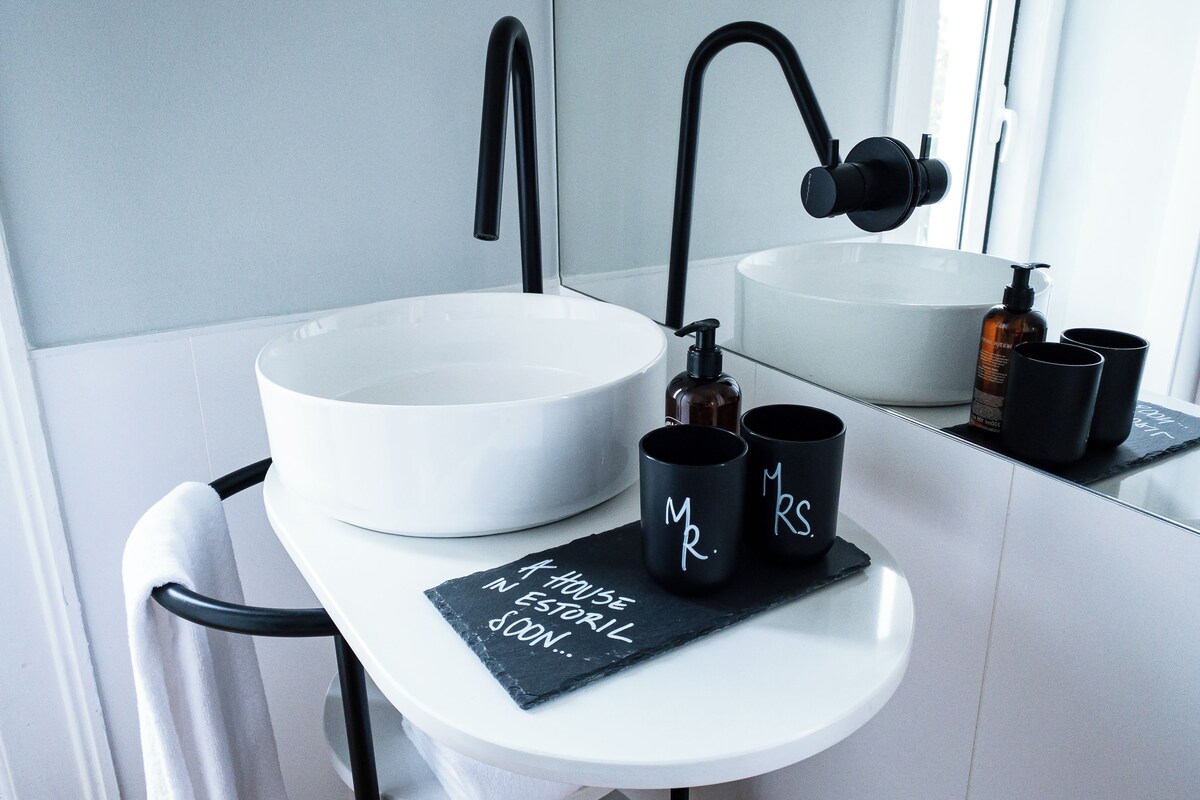 A modern bathroom with a white sink, black faucet, and two black mugs with 'Mr.' and 'Ms.' on them.