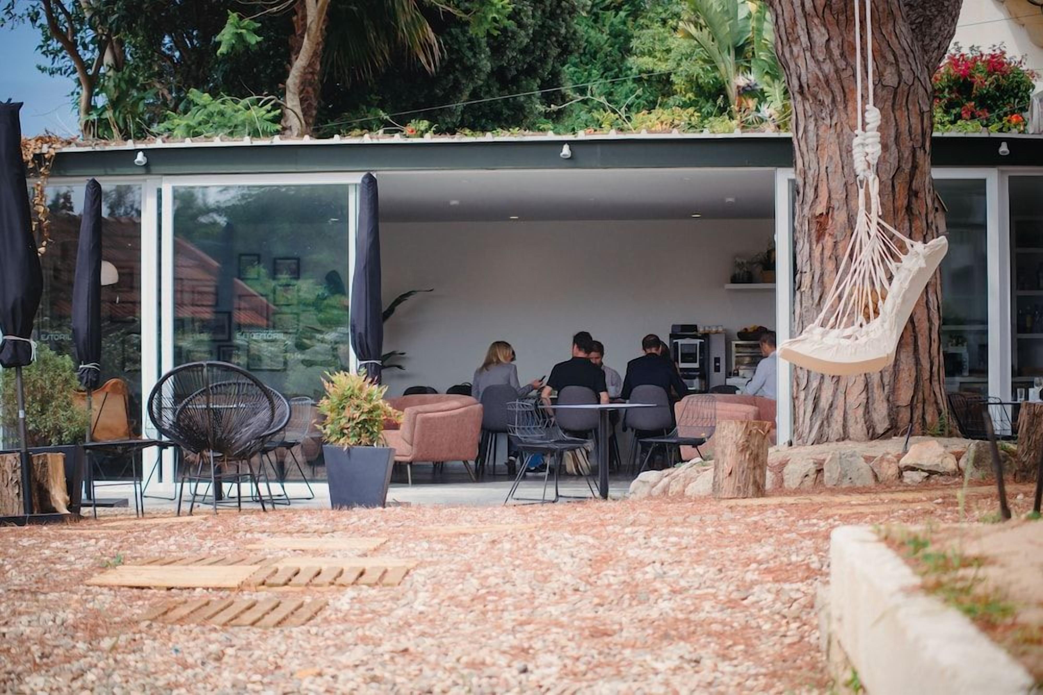 Un espacio al aire libre con mesas, sillas, una hamaca y el tronco de un árbol.