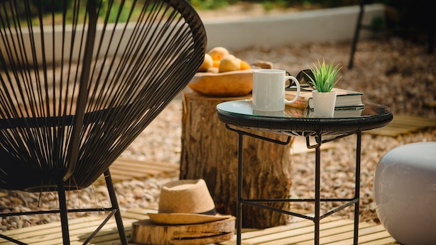 Une chaise en osier, une petite table ronde avec une tasse blanche et une plante en pot, et un tabouret en bois sur lequel sont posés un bol de pain et un chapeau.
