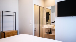Une chambre moderne comprenant un lit, une table de chevet en bois, une armoire, un téléviseur intégré et une salle de bain avec un lavabo et un miroir.