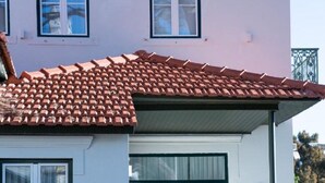 Un bâtiment à deux étages, doté d’un toit recouvert de tuiles rouges, de murs blancs et de grandes fenêtres. On y trouve un petit espace extérieur aménagé avec une chaise et un pot de fleurs décoratif.