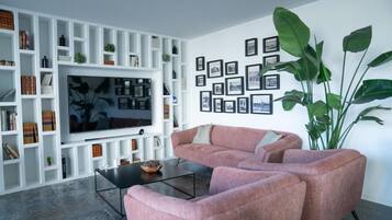 A modern living room with a large flat-screen TV, a pink sectional sofa, a coffee table, and a white shelving unit filled with books and decorative items.