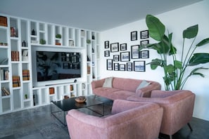 A modern living room with a large flat-screen TV, a pink sectional sofa, a coffee table, and a white shelving unit filled with books and decorative items. - A House In Estoril (Cascais)
