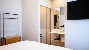 Une chambre moderne comprenant un lit, une table de chevet en bois, une armoire, un téléviseur intégré et une salle de bain avec un lavabo et un miroir.