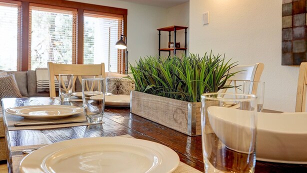 Een eetruimte met een houten tafel voor vier personen, omringd door stoelen. In het midden staat een grote potplant, en op de achtergrond een plankje met een beeldje van een kat.