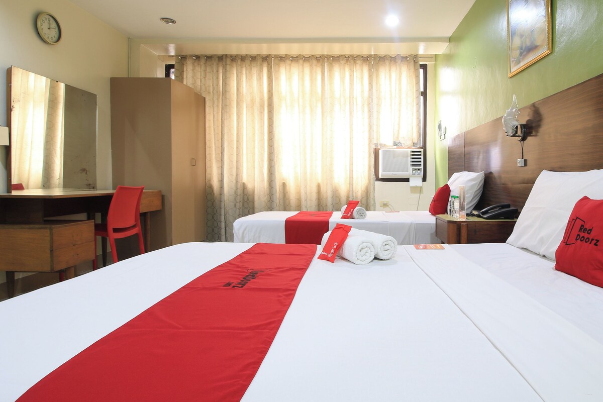 A hotel room with a bed, a desk, a chair, and a nightstand with a phone and a bottle.