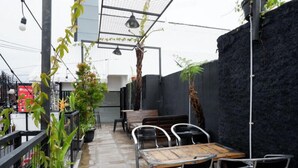 Un patio al aire libre con una mesa de madera y sillas de metal, rodeado de plantas en macetas y un muro negro.
