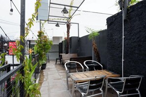 Un patio al aire libre con una mesa de madera y sillas de metal, rodeado de plantas en macetas y un muro negro.