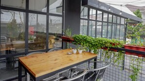 A wooden table with metal chairs, a hanging plant, and a view of the street through large windows. - Reddoorz @ Demangan Sari Residence (Depok)