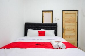 Una cama bien hecha con cabecera negra, ropa blanca de cama y una colcha roja. Se observan dos puertas de madera al fondo.