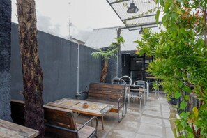 Un patio al aire libre con mesa y sillas de madera, rodeado de follaje y una cerca de metal.