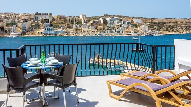 A balcony with a table set for dining, overlooking a coastal cityscape with buildings and boats. - GetawaysMalta - Harbour Lights Seafront Penthouse with Terrace St Paul's Bay (St Paul's Bay)