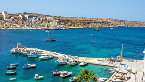 A coastal marina with various boats docked, a clear blue sea, and a hillside with buildings in the background. - GetawaysMalta - Harbour Lights Seafront Penthouse with Terrace St Paul's Bay (St Paul's Bay)