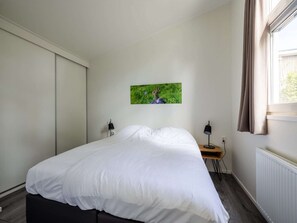 Een slaapkamer met een bed, nachtkastje, lamp, foto aan de muur en een raam met gordijnen.