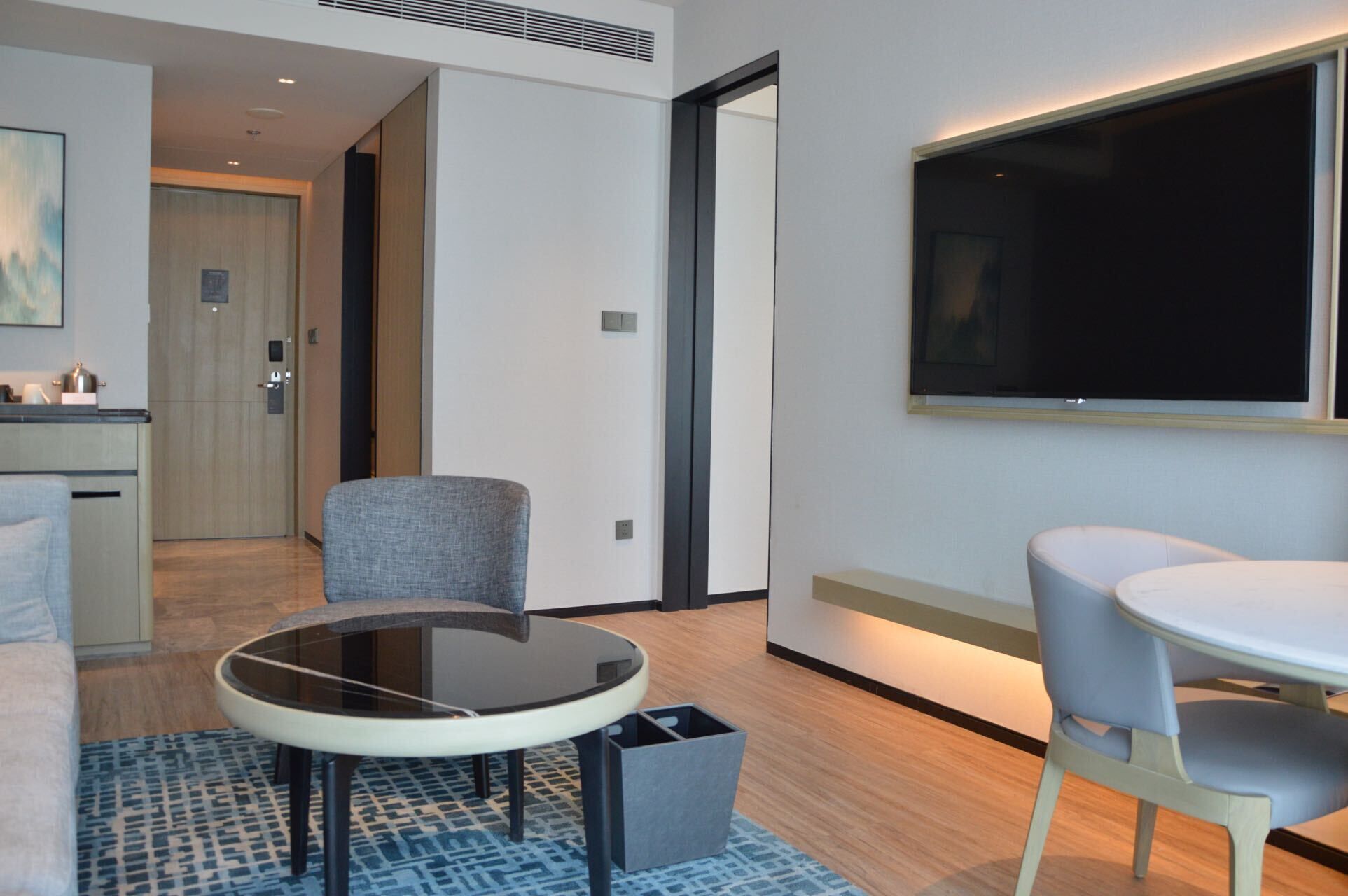 A modern hotel room with a round glass table, two chairs, a flat-screen TV, and a kitchenette in the background.