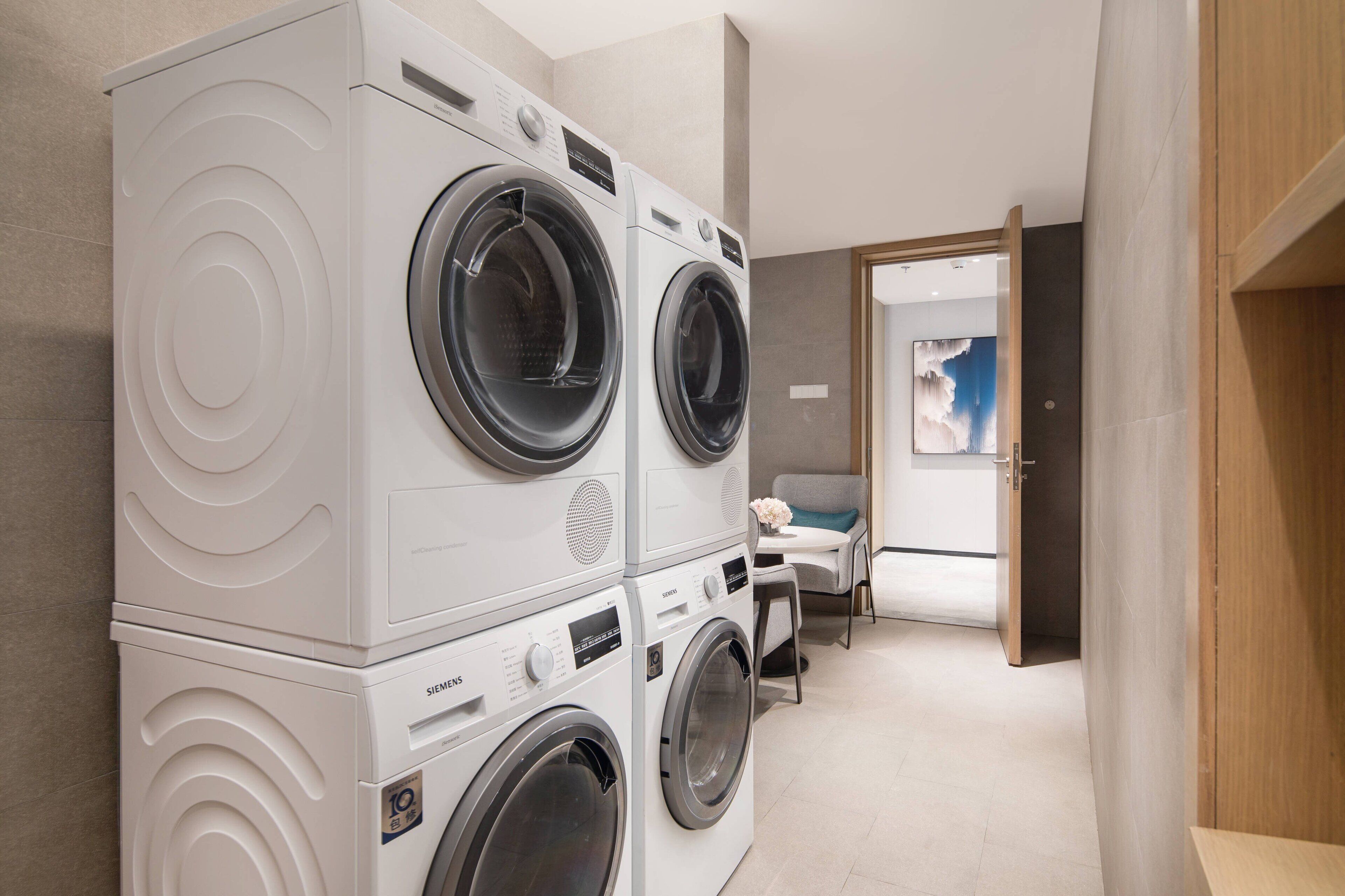 A modern laundry room with stacked front-loading washers and dryers, a small seating area with a table and chairs, and a door leading to another room.