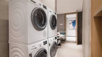 A modern laundry room with stacked front-loading washers and dryers, a small seating area with a table and chairs, and a door leading to another room.