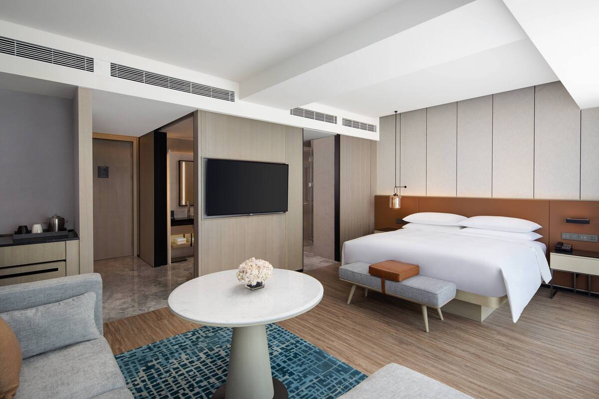 Ein modernes Hotelzimmer mit einem großen Bett, einem Flachbildfernseher, einem kleinen Tisch mit einer Vase sowie einer Sitzecke mit Sofa und Sessel.