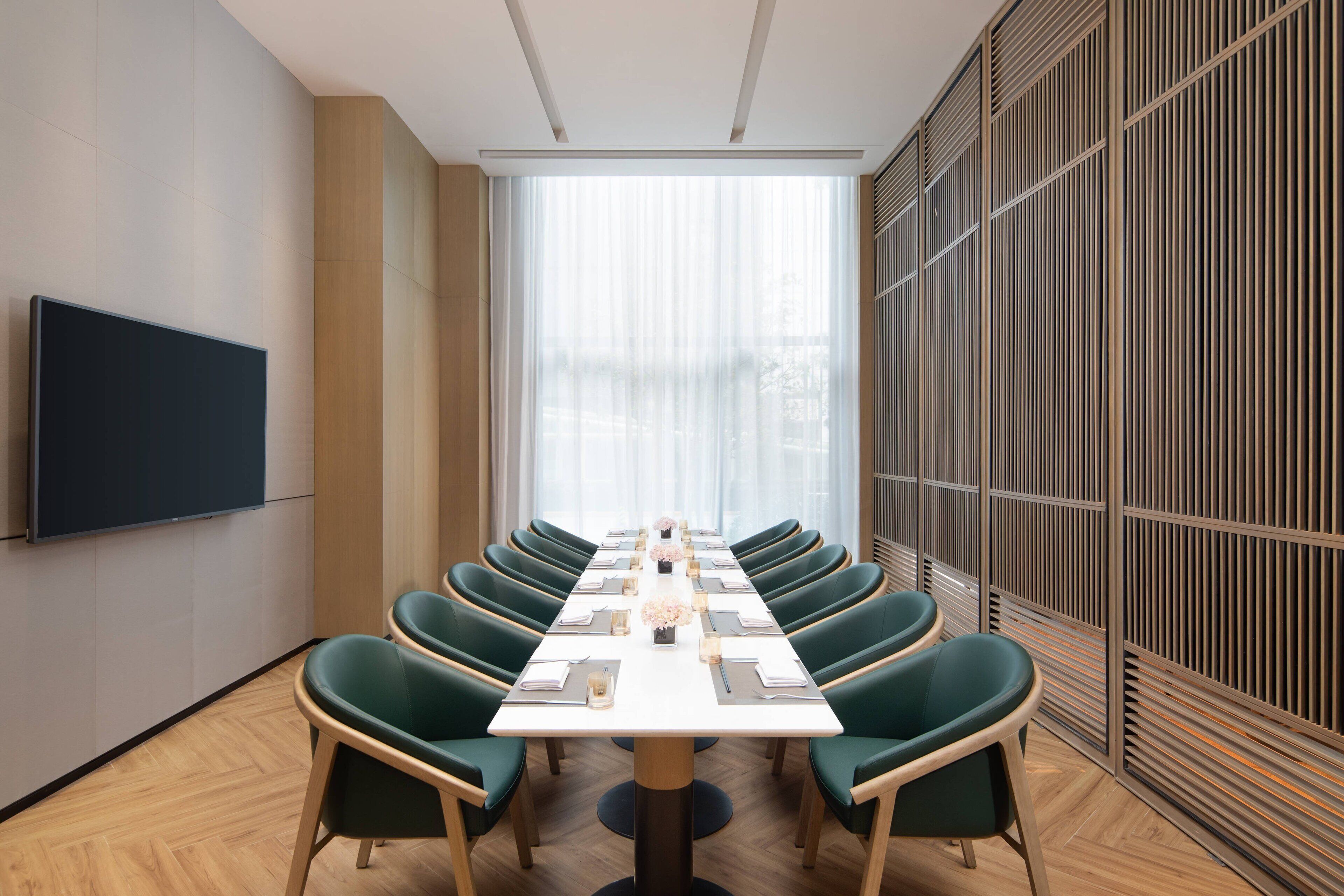 Un salón de conferencias con una mesa larga, sillas verdes, un televisor de pantalla plana y un amplio ventanal con cortinas translúcidas.