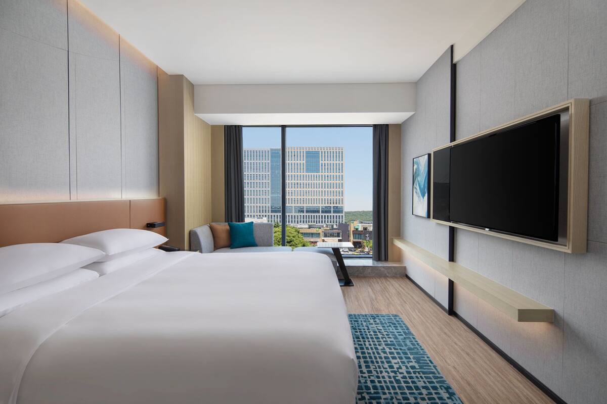 Ein modernes Hotelzimmer mit einem großen Bett, einem flachbildfernseher an der Wand und Blick auf die Stadt durch große Fenster.