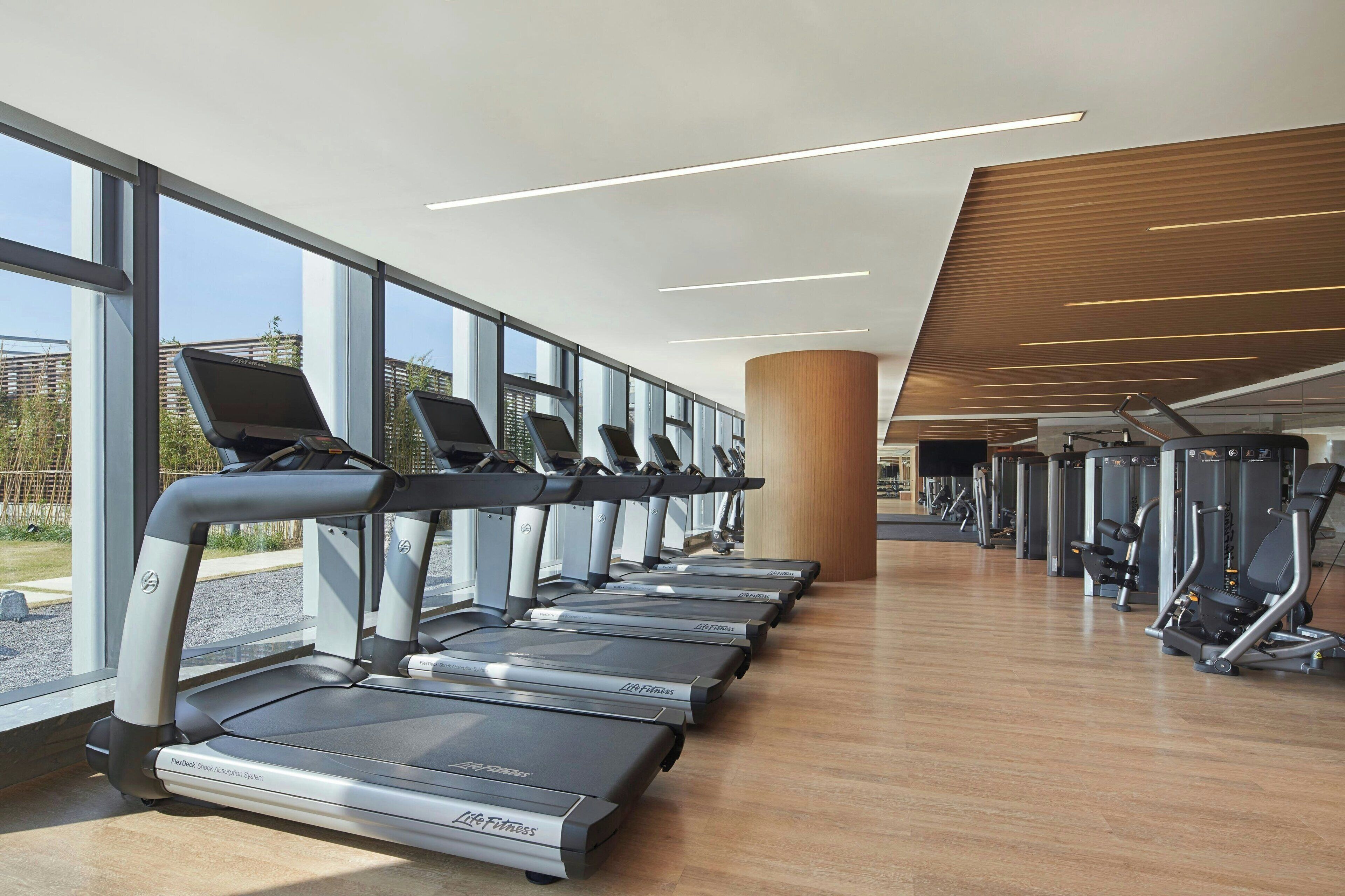 Une salle de sport avec des tapis de course alignés le long d’un mur doté de grandes fenêtres, un pilier en bois et du matériel d’exercice.