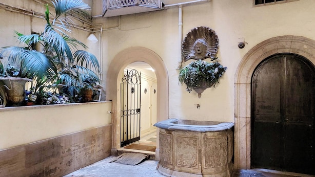 A narrow alley with a stone fountain, a large potted plant, and a closed door. - Campo de Fiori, Navona - Fashion Domus (Roma)