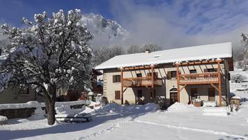 Un bâtiment enneigé, doté de balcons en bois, d’un mur de pierre et d’un arbre recouvert de neige devant.